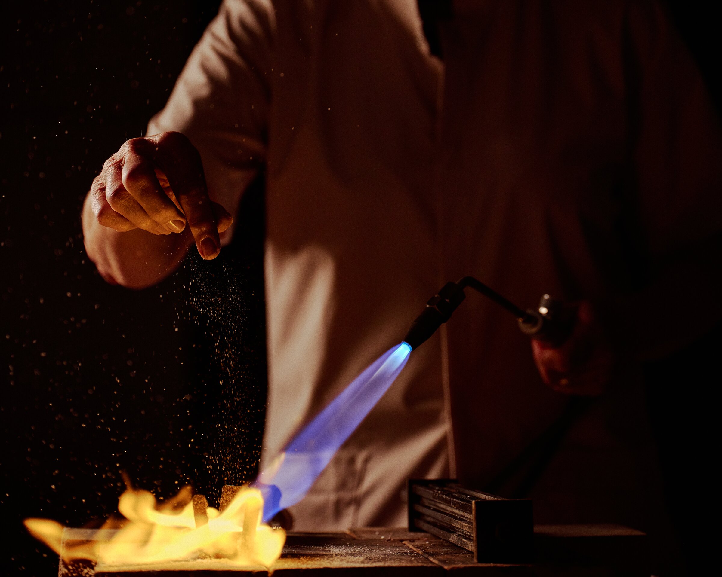 Goldsmith sprinkling flux over a torch flame at the workbench