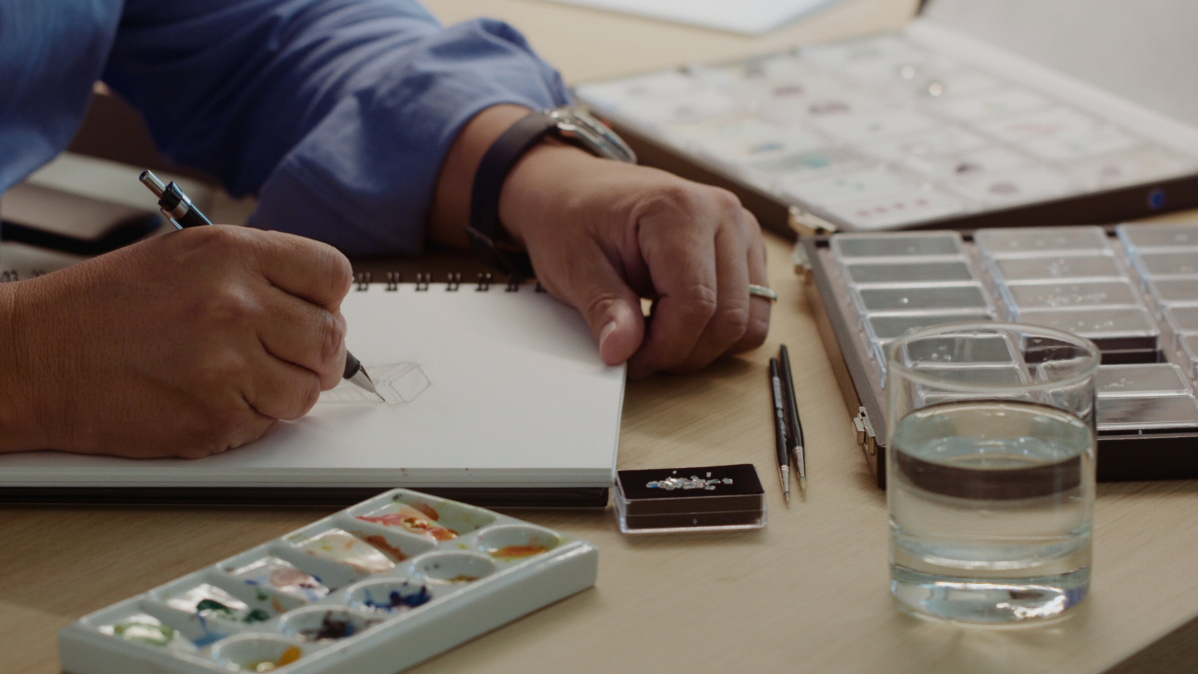 Jewellery designer sketching a ring, watercolour palette and loose stones nearby