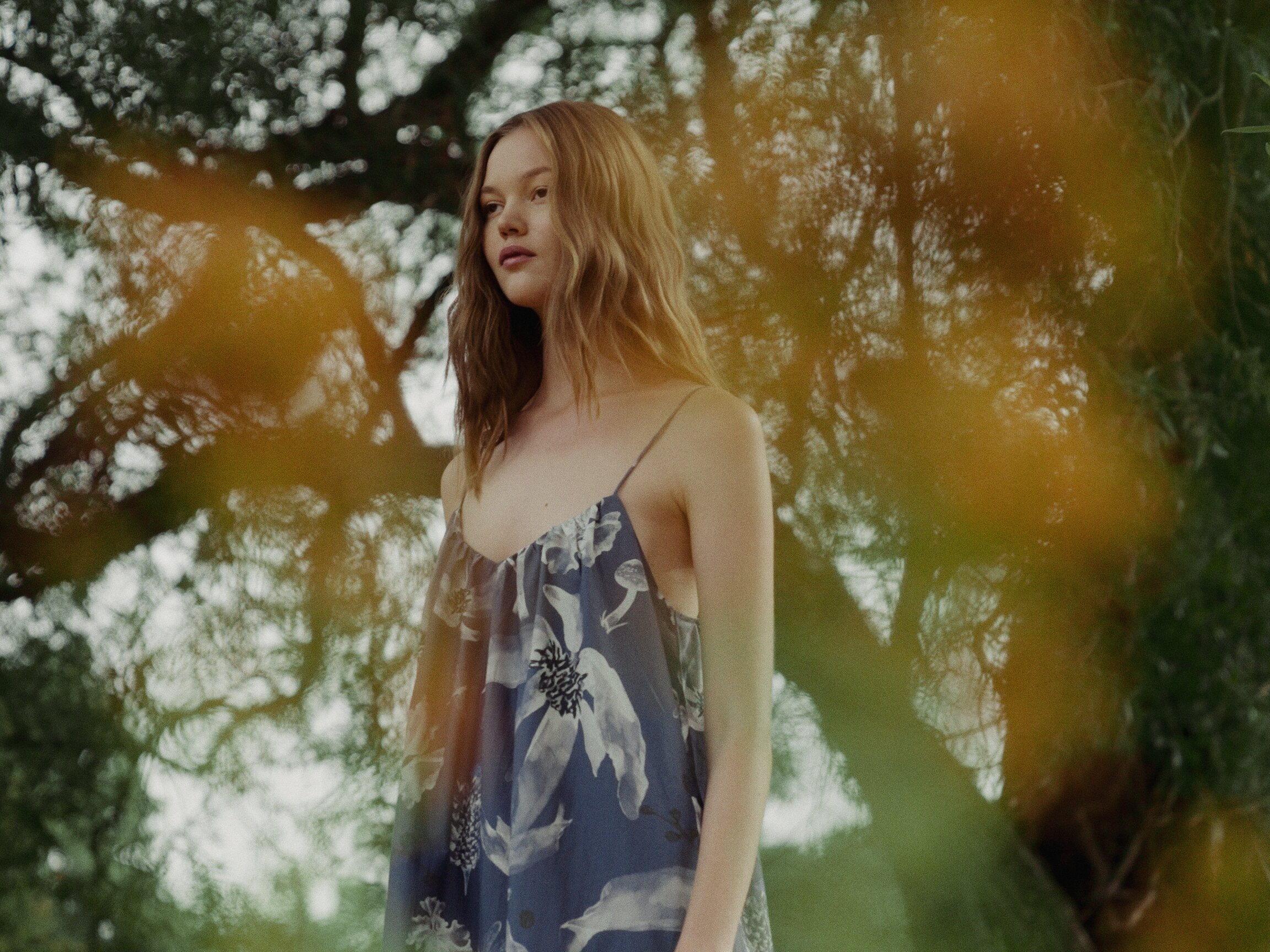 Model in a blue floral slip dress, portrait through backlit foliage