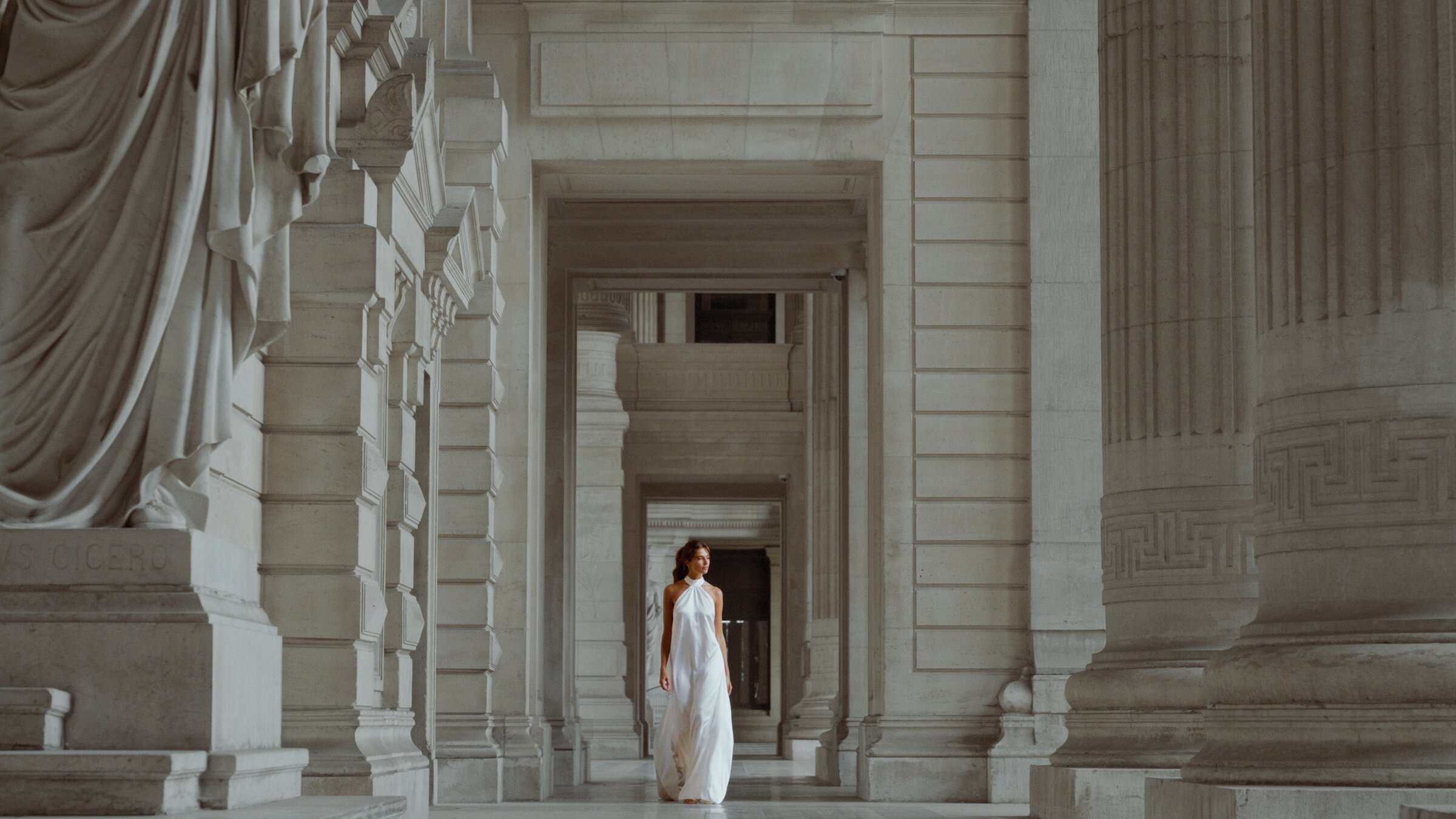 Model in a white halter gown walking through a monumental stone colonnade