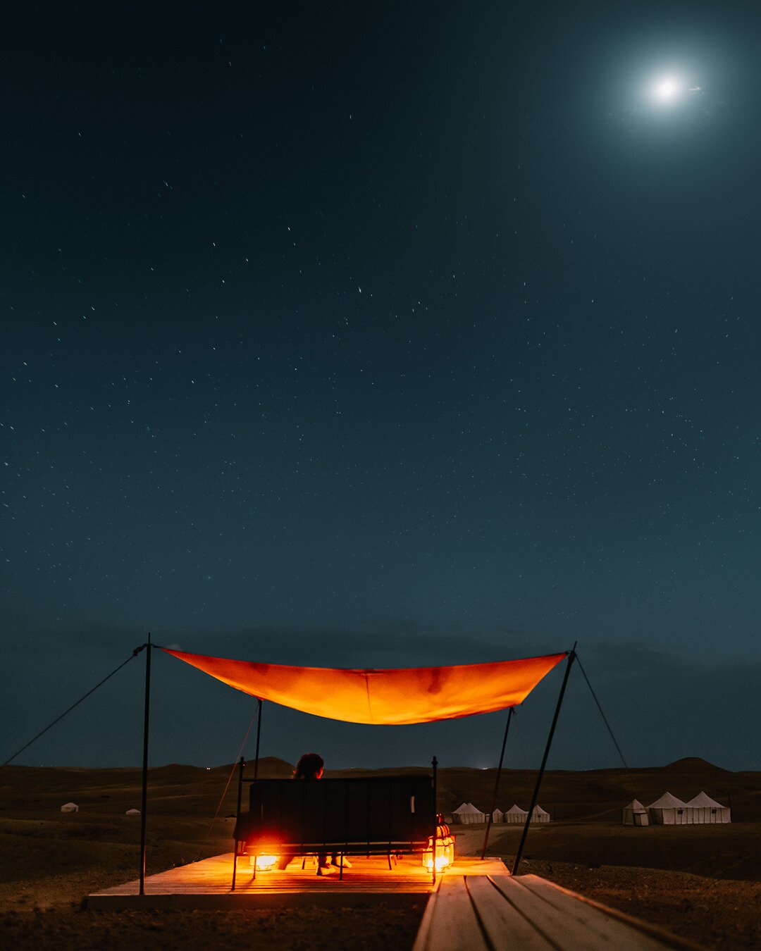 Desert camp at night, lantern light under a canopy and a starlit sky